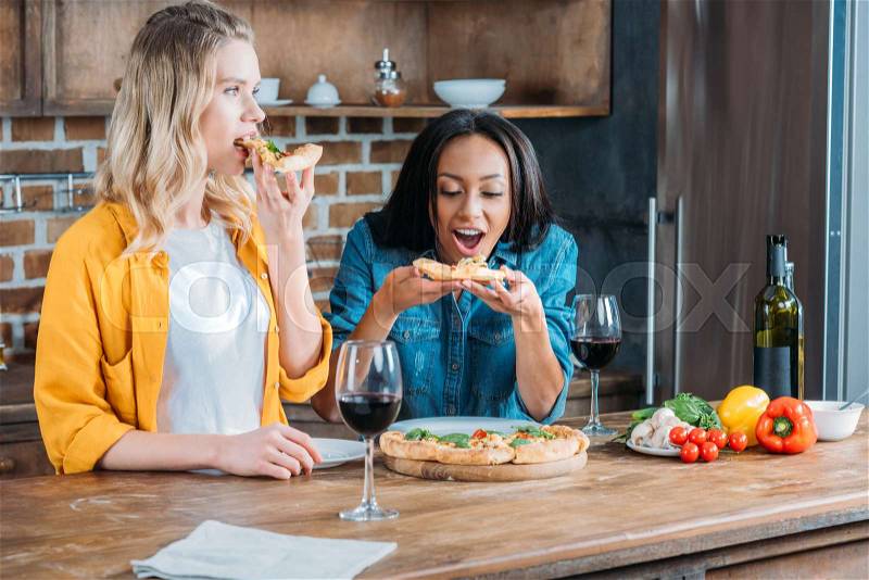 Cheerful multiethnic women eating pizza and drinking wine at home, stock photo