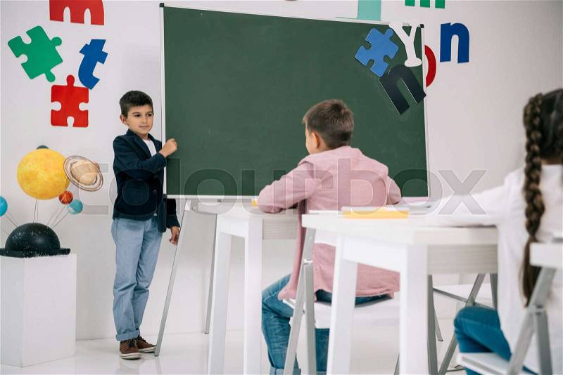 Schoolboy standing at chalkboard and looking at classmates sitting at desks, stock photo