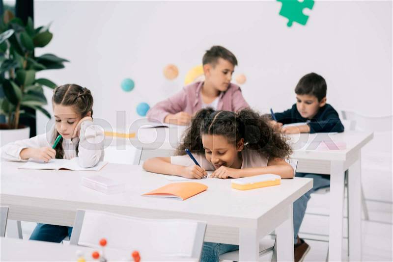 Multiethnic group of schoolkids writing in exercise books and studying together in classroom, stock photo
