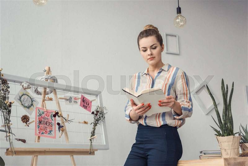 Concentrated beautiful female florist reading book at workplace, stock photo