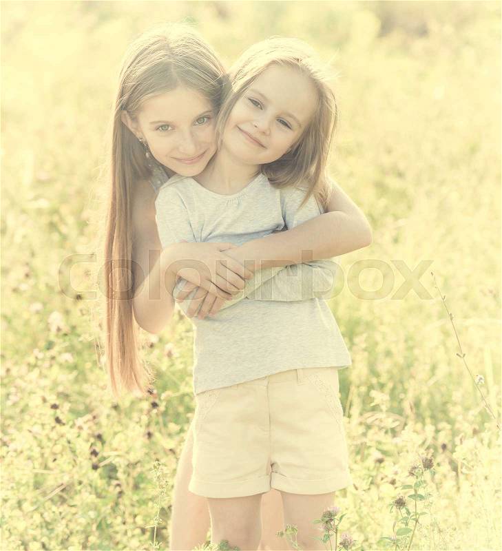 Cute sibling sisters hugging each other, standing in the field with chamomile flowers, stock photo
