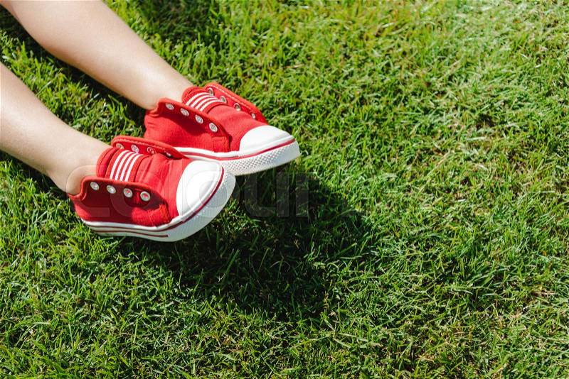 Close-up partial view of little feet in red sneakers on green grass, stock photo