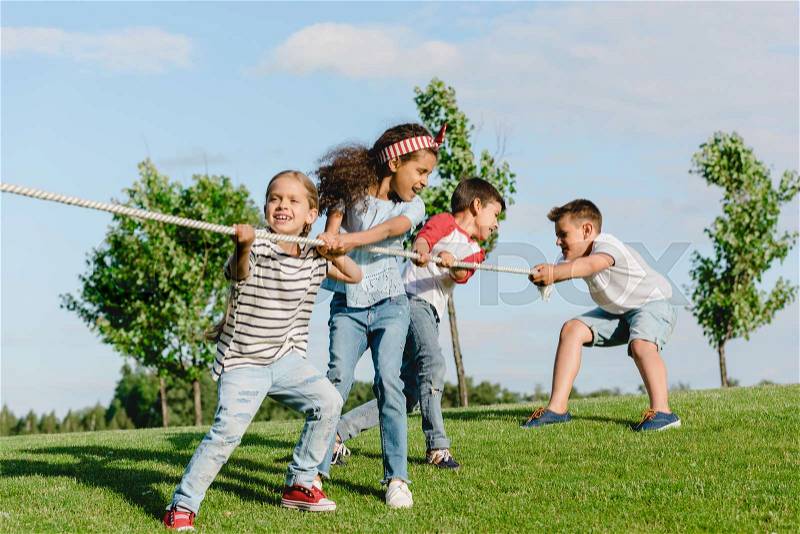 Cheerful multiethnic kids pulling rope and playing tug of war in park, stock photo