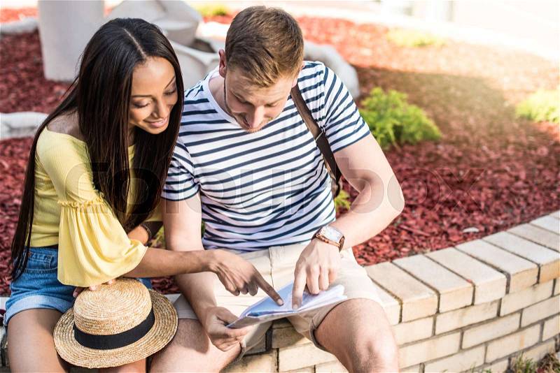 Cheerful young multiethnic couple of tourists pointing at map while sitting outdoors , stock photo