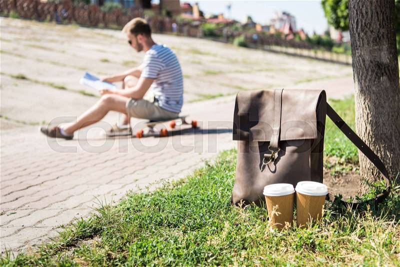Close-up view of brown leather shoulder bag with disposable coffee cups and man sitting on skateboard behind, stock photo