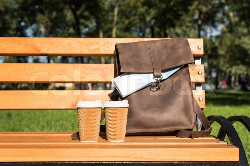 Close-up view of brown leather bag with map and disposable coffee cups on wooden bench, stock photo