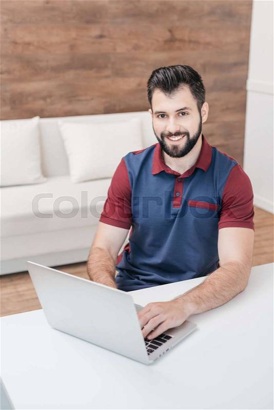 Handsome bearded man using laptop and smiling at camera at home, stock photo