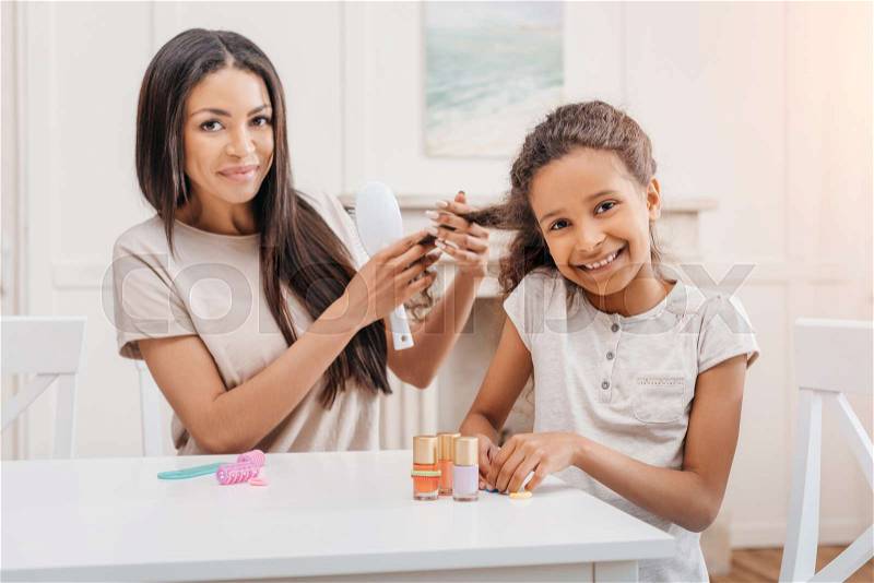 Happy african american daughter doing manicure while mother combing her hair, stock photo