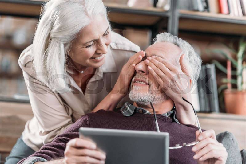 Woman covering eyes of Senior man with tablet at home, stock photo