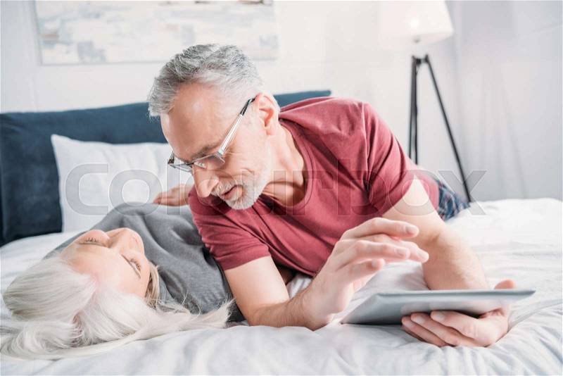 Happy senior couple with digital tablet resting in bed together, stock photo