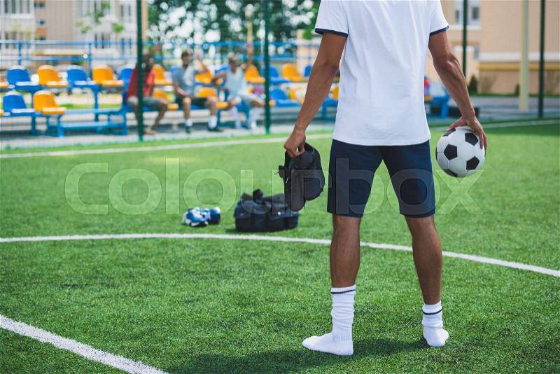 Back view of soccer player with ball and football boots in hands standing on pitch, stock photo