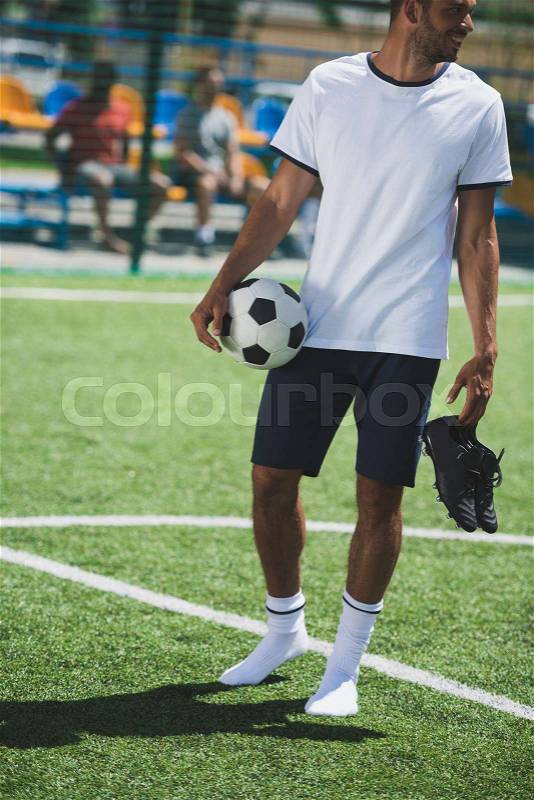 Athletic soccer player holding ball and boots in hands while standing on pitch, stock photo