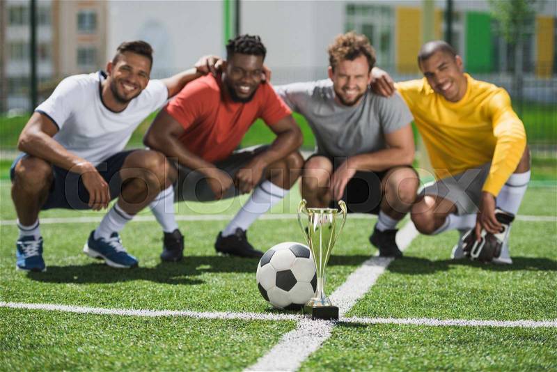 Smiling multiethnic soccer team with goblet on soccer pitch after game, stock photo