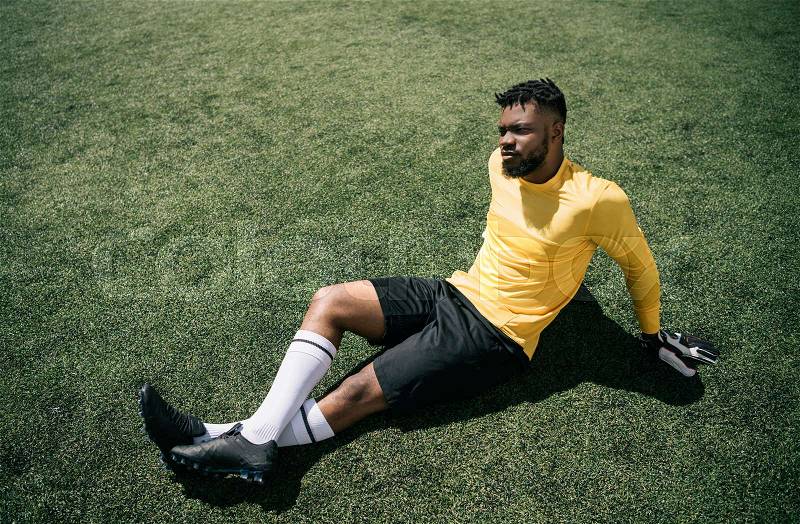 Athletic african american goalkeeper sitting on soccer pitch, stock photo