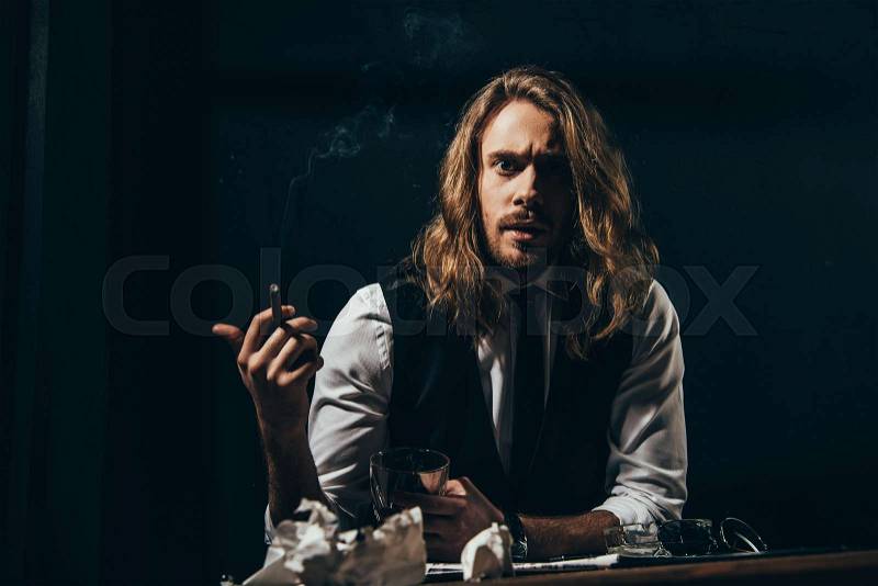 Bearded handsome long haired man in formal wear smoking cigar while holding glass of whisky and looking at camera, stock photo