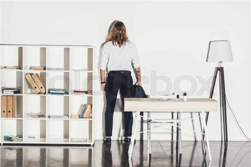 Back view of young businessman in formal wear standing near wall in office , stock photo