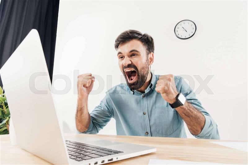 Portrait of excited businessman screaming and looking at laptop screen at workplace in office, stock photo