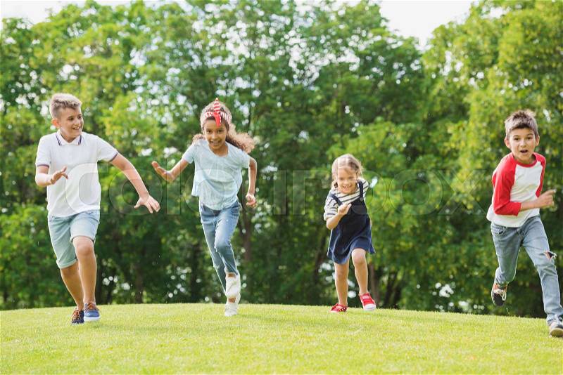 Adorable happy multiethnic kids playing and running together on green grass in park, stock photo