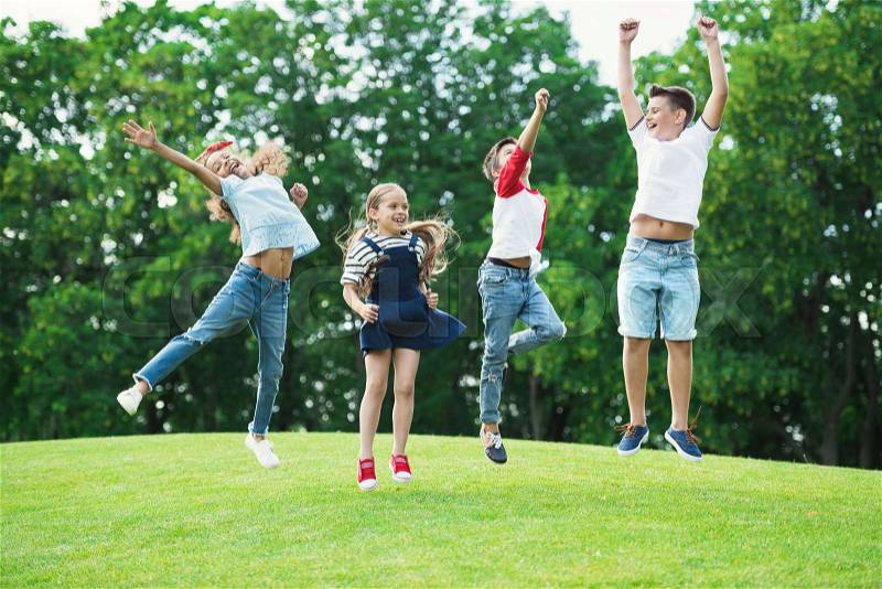 Adorable happy multiethnic kids playing and jumping on green meadow in park, stock photo