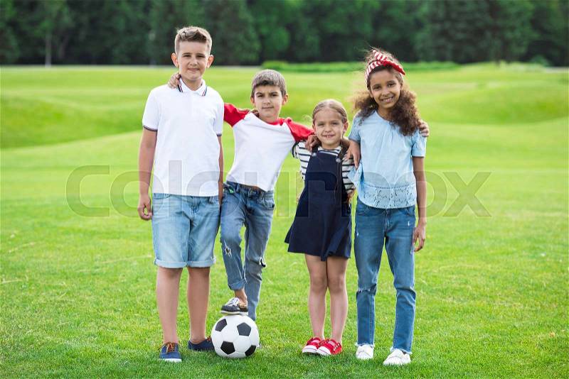 Adorable multiethnic kids standing with soccer ball and smiling at camera in park, stock photo