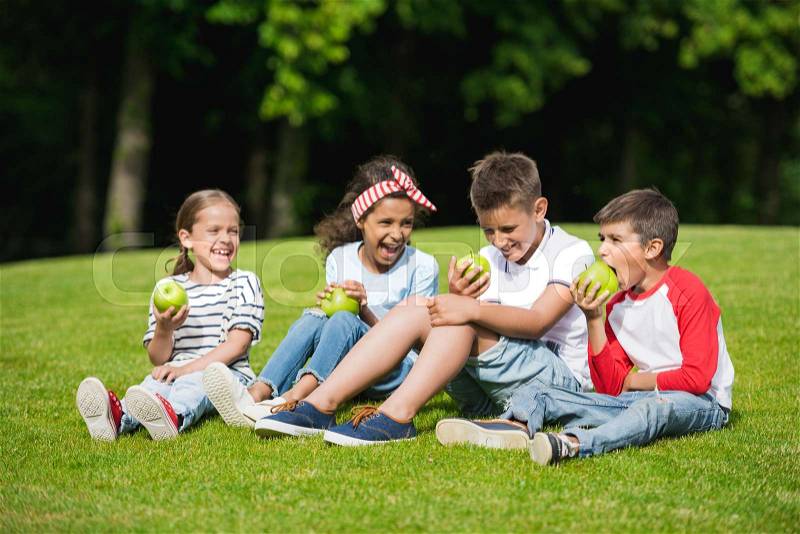 Happy multiethnic children eating green apples while sitting together on green grass, stock photo