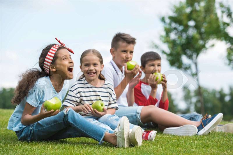Happy multiethnic children eating green apples while sitting together on green grass, stock photo