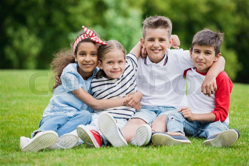 Happy multiethnic kids sitting embracing and smiling at camera in park, stock photo