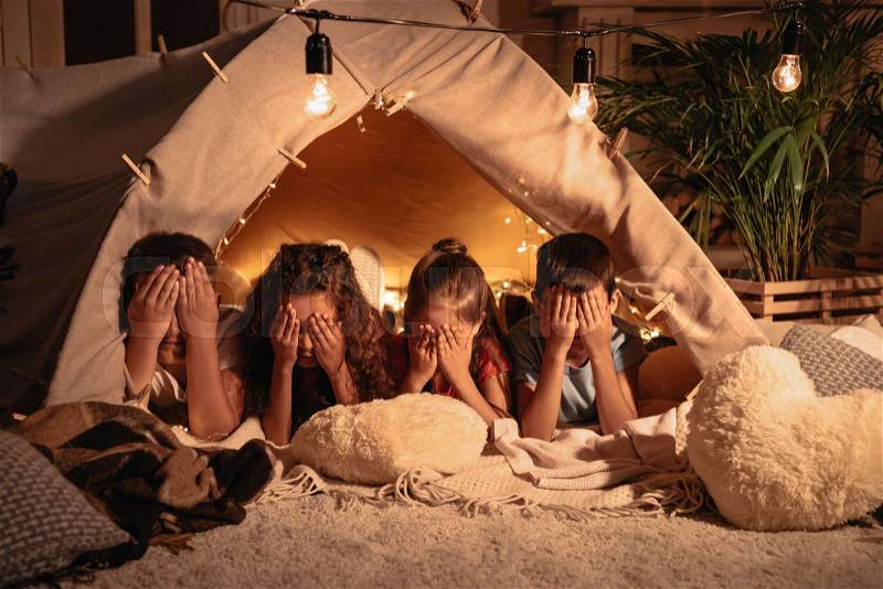 Group of children covering face with hands while resting in tent at home, stock photo