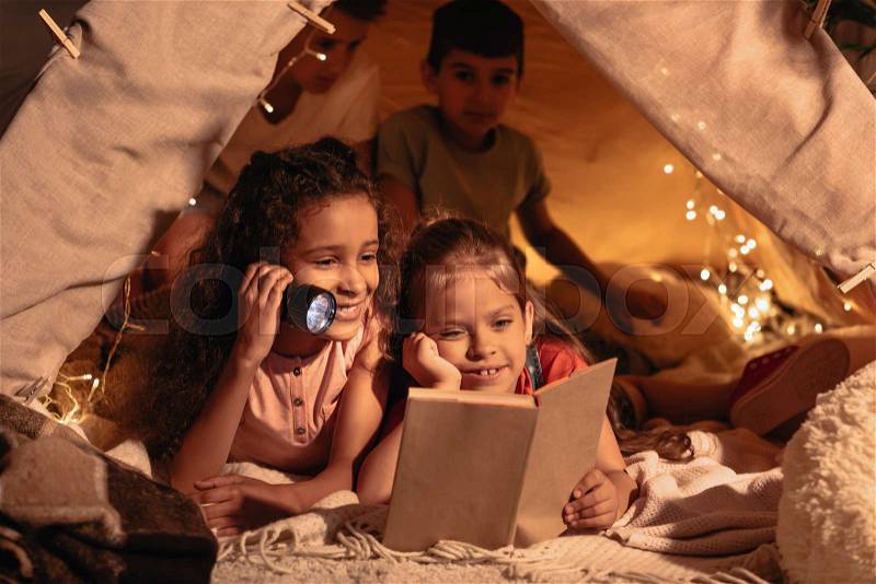 Smiling girls with lighter reading book in handmade tent at home, stock photo