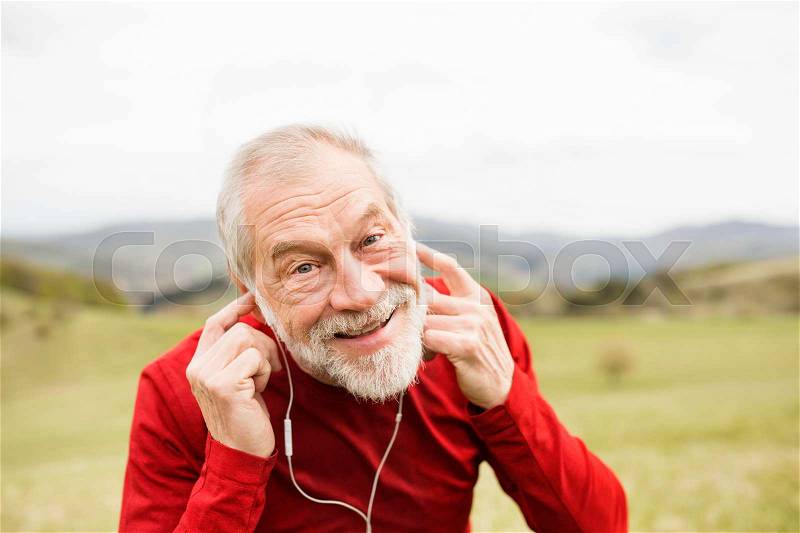 Active senior runner in red sweatshirt with earphones listening music outside on green hills, taking break, resting, stock photo