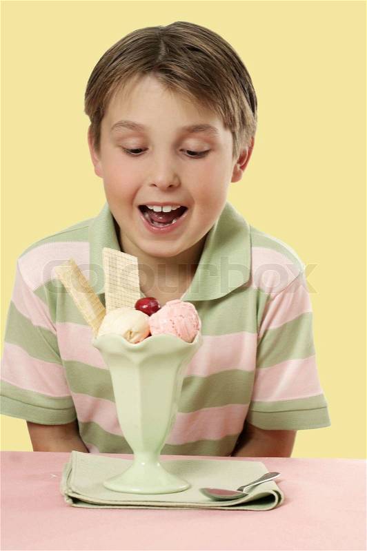 Boy looks at an ice cream sundae with wafers and cherry decoration placed on a table, stock photo
