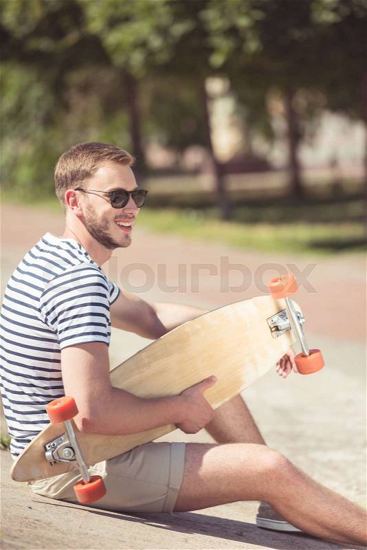 Happy handsome man in sunglasses sitting and holding skateboard, stock photo