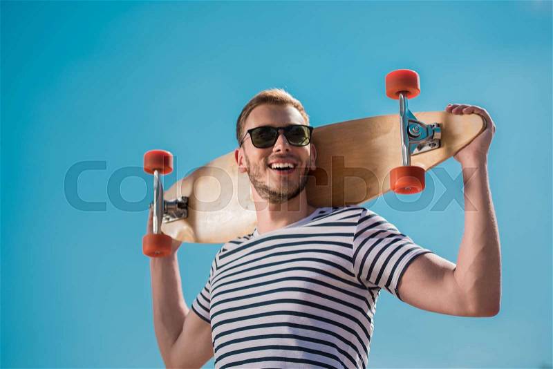 Happy handsome man in sunglasses holding skateboard, blue sky on background, stock photo