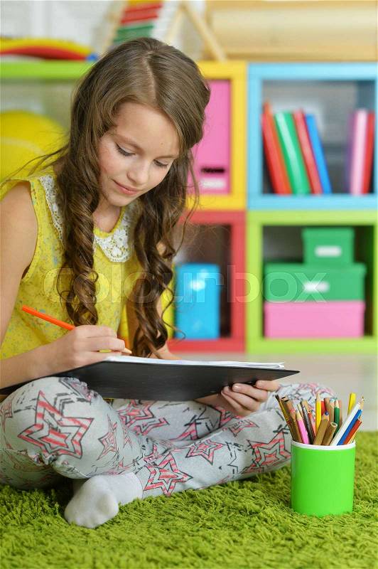 Little girl painting with pencil in her room with bookshelves on background, stock photo