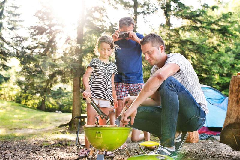 Beautiful family enjoying camping holiday in forest. Father cooking meat on barbecue grill, grandfather taking picture with camera, stock photo