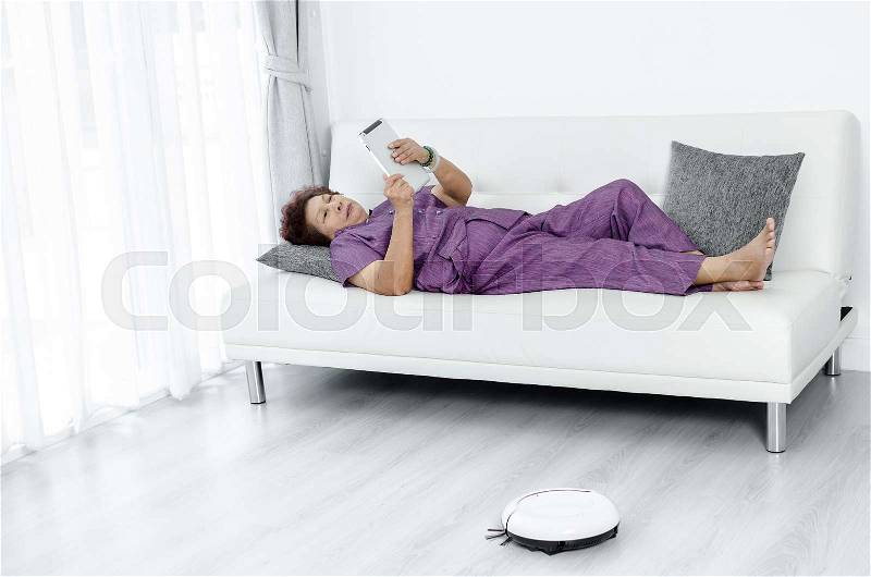 Asian senior woman lying on couch and using tablet. Robot vacuum cleaner runs in the room, stock photo