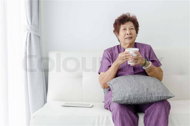 Asian senior woman drinking water,sitting on couch near window at home, stock photo