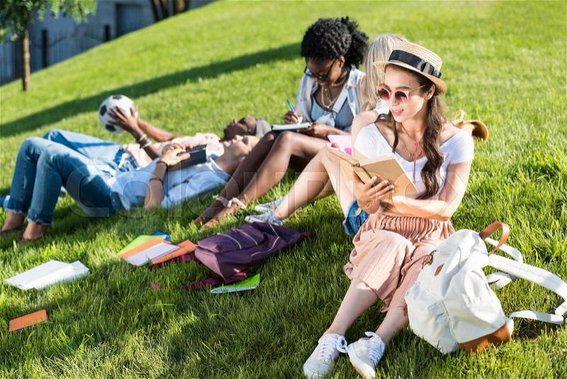 Multiethnic group of students studying and resting on green grass in park, stock photo