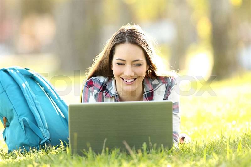 Front view of a happy student watching on line content in a laptop lying on the grass of a park, stock photo