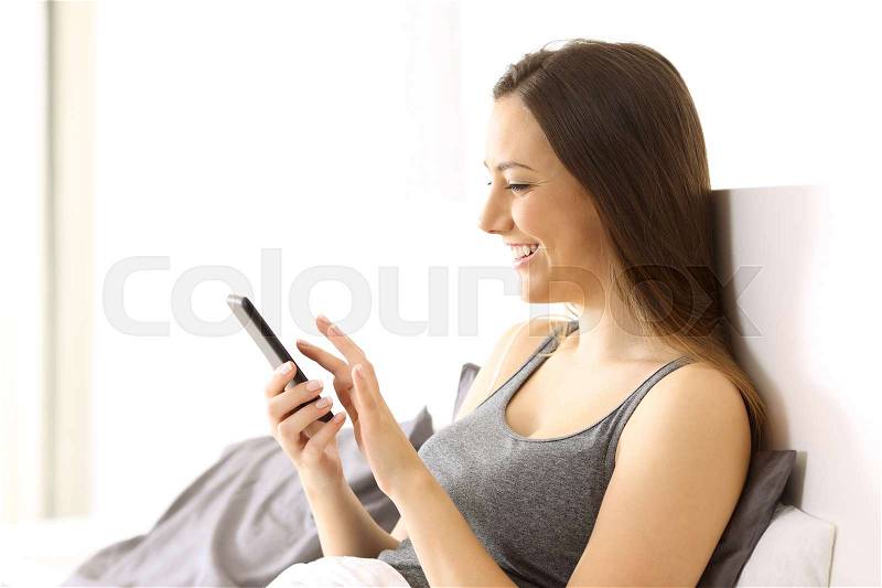 Side view of a woman spending free time using a smart phone on the bed of an hotel room or home, stock photo