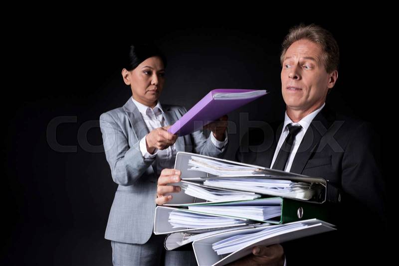 Frustrated mature businessman holding pile of folders while female colleague putting folder on the top isolated on black, stock photo