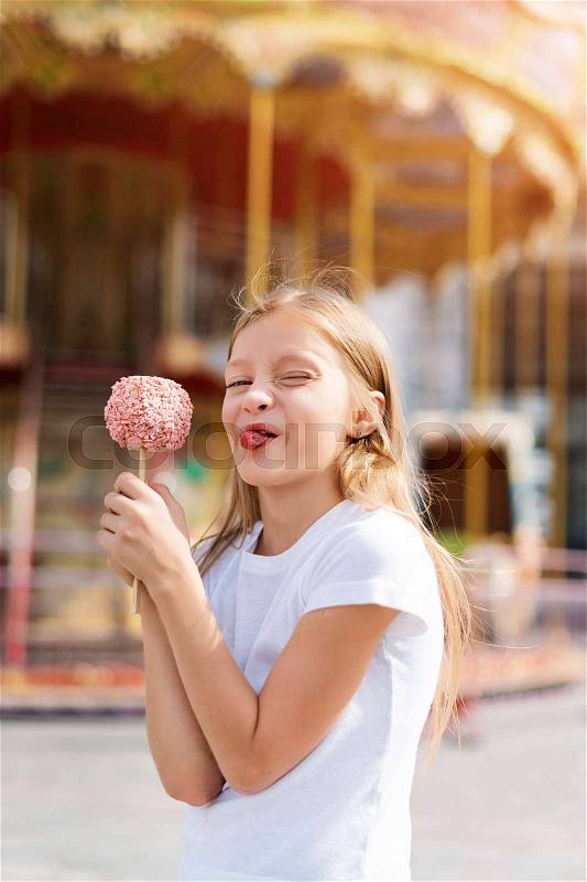 Cute little girl eating candy apple and posing at fair in amusement park, stock photo