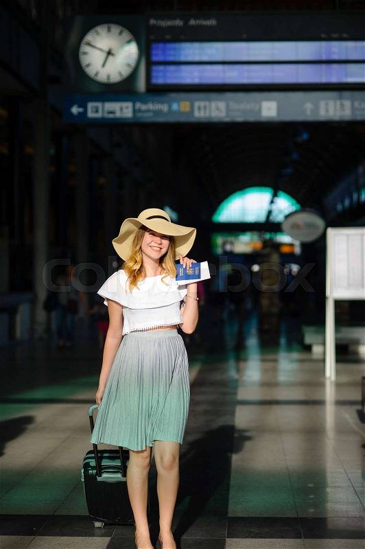 Charming girl in sun-hat in the waiting room of the train station. Nearby is a suitcase. In hand her passport and tickets. Time of summer holidays, stock photo