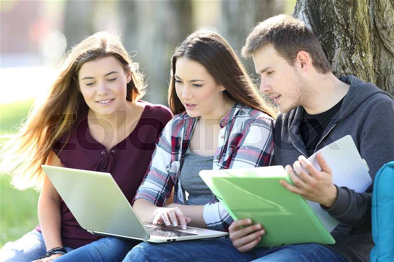 Three attentive students learning on line together with a laptop and notebook sitting on the grass in a park, stock photo