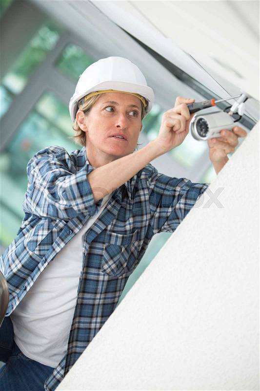Female technician installing camera on wall using electric cordless drill, stock photo