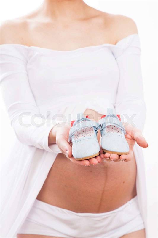 Pregnant woman in a white dress holding a tiny baby shoes, stock photo