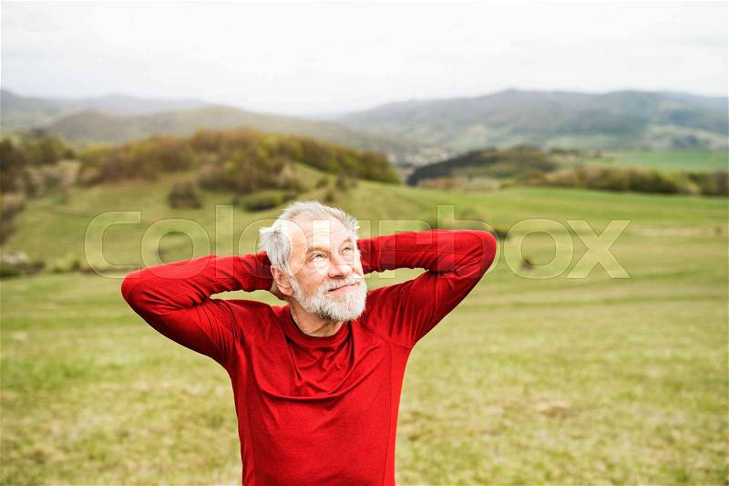 Active senior runner in red sweatshirt outside on green hills, taking break, resting, stock photo