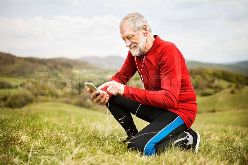 Active senior runner outside on green hills with smart phone and earphones, resting, listening music, stock photo