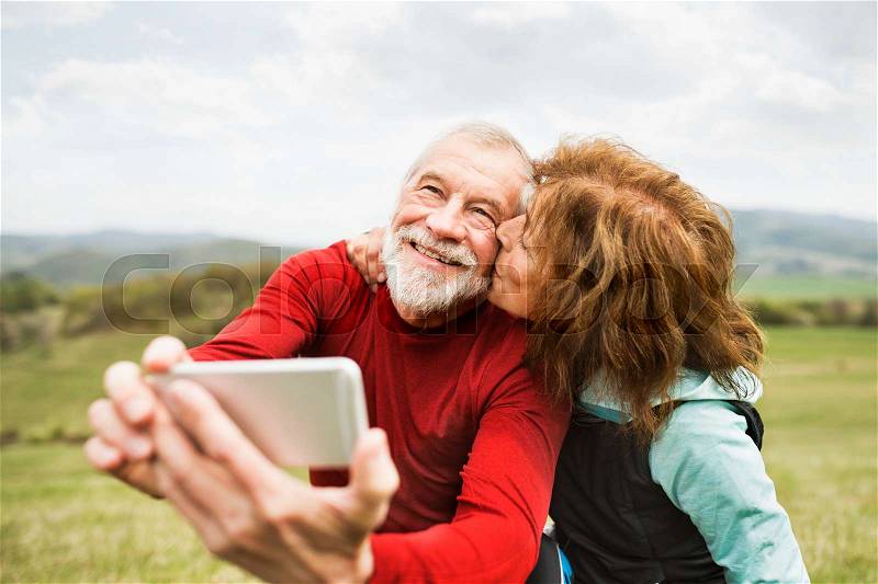 Active senior runners outside on the green hills taking photo with smart phone. Woman kissing man on cheek. Calvary in Banska Stiavnica, Slovakia, stock photo