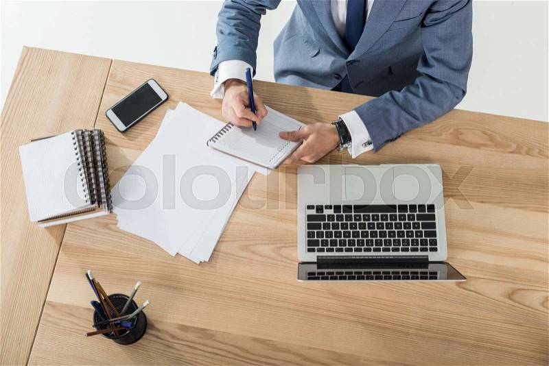 Cropped shot of businessman writing in notebook at workplace with laptop and papers in office, stock photo